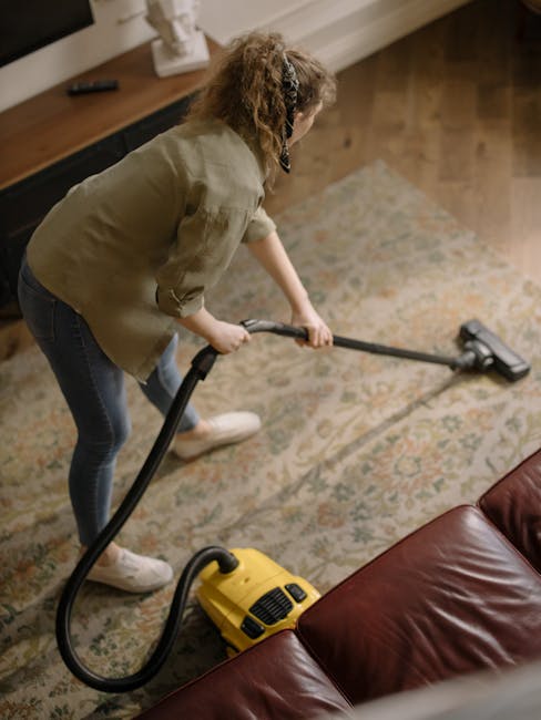 Close-up of a person vacuuming a patterned carpet in a living room, with the vacuum cleaner's head prominently in focus. The carpet features a floral design with muted colors, and the vacuum hose exte