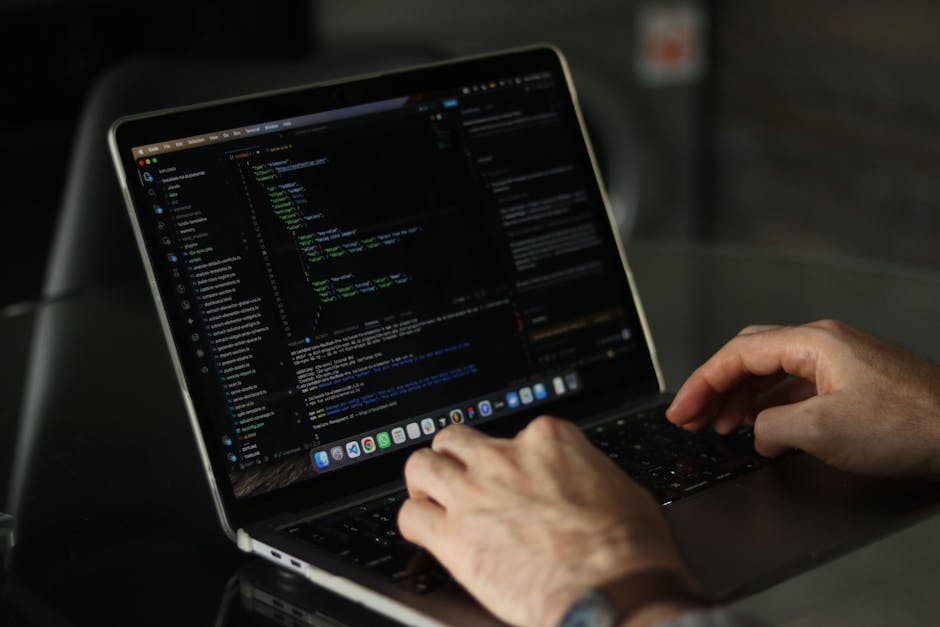 Close-up of a person's hands typing on a laptop with a black keyboard, displaying lines of code and programming interfaces on the screen. The laptop is placed on a dark desk in a well-lit room with a