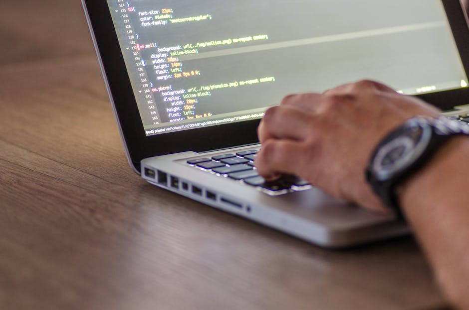 Close-up of a person's hands typing on a laptop with a black keyboard, displaying lines of code and programming interfaces on the screen. The laptop is placed on a dark desk in a well-lit room with a